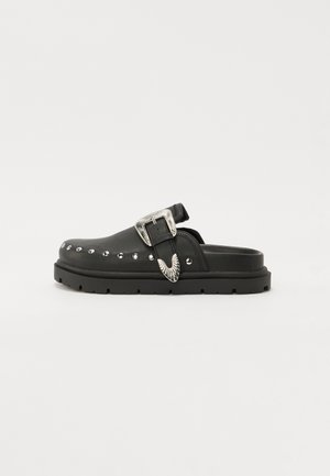 Sabot en cuir noir avec une semelle épaisse et texturée, boucle décorative en argent et accents cloutés sur le côté.