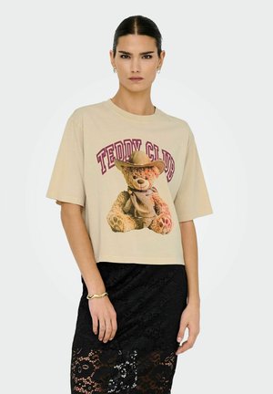 Beige cropped t-shirt med grafik af en bamse i cowboyhat. Parret med en sort blonde-nederdel, der viser struktur og mønster.