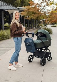 Zelený kočárek s kapucí, připevněný k tmavě modré tašce na plenky. Uživatel má na sobě hnědý semišový kabát a modré džíny, drží kelímek na kávu.