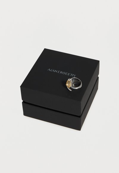 Anillo de plata con una piedra oval dorada colocado sobre una caja de joyas negra de Nolan Croletti contra un fondo blanco.