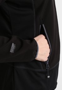Chaqueta softshell negra con bolsillo con cremallera, costuras contrastantes y una etiqueta ICETECH en la manga. Presenta un tejido suave y texturizado.