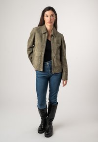 Giacca in suede verde oliva con colletto, dotata di due tasche frontali e chiusura con zip. Indossata sopra un top nero e jeans blu, con stivali neri.