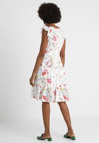 Frau mit Afro-Frisur, gekleidet in einem weißen, knielangen Kleid mit floralen Mustern und Rüschensäumen sowie grünen, offenen, flachen Schuhen mit niedrigen Absätzen, die mit dem Rücken zur Kamera steht.