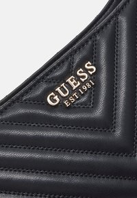Cuir matelassé noir avec une couture inclinée et un logo métallique doré « GUESS EST 1981 » dans le coin.