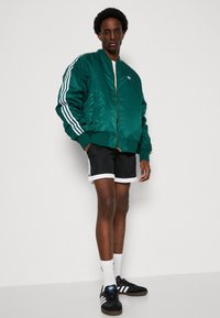 Groene satijnen bomberjack met witte strepen op de mouwen, zwarte shorts, witte crew-sokken en zwarte sneakers met witte accenten.