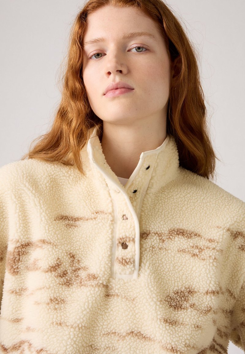 Beiger Fleece-Pullover mit strukturierter Oberfläche, braunem abstraktem Muster und hohem Kragen. Mit Knopfverschluss und kontrastierendem Besatz.
