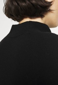 Cheveux courts brun foncé au-dessus de l'encolure d'un pull noir côtelé à col montant, sur une personne de dos.