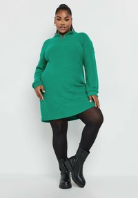 Vestido de punto verde con cuello con cremallera, mangas largas y corte relajado, combinado con botas altas negras de textura.