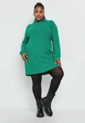 Robe pull verte côtelée avec un col zippé, des manches longues et une coupe décontractée, associée à des bottes noirs texturées jusqu'aux genoux.