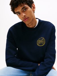 Felpa blu navy con logo dorato e design a alloro. Realizzata in morbido tessuto, presenta un colletto rotondo e polsini a coste. Indossata con jeans azzurri.