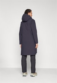 Lungo cappotto blu navy con cappuccio, realizzato in tessuto morbido. Presenta una linea dritta e maniche lunghe. Abbinato a pantaloni grigi e sneakers chiare.