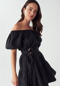 Melnā off-the-shoulder kleita ar pūkainām piedurknēm, savilktā jostasvietā ar zelta apaļu sprādzi un plūstošu, slāņotu svārku.