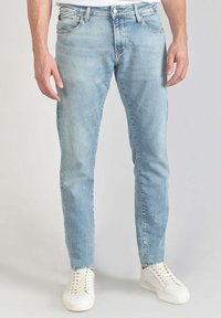 Vaqueros de mezclilla azul claro con corte slim, textura deslavada, cinco bolsillos, pasadores para cinturón y cierre con cremallera, combinados con zapatillas blancas.