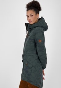 Parka verde a fantasia mimetica con cappuccio, dotata di zip frontale, regolazioni con cordino e dettagli in pelle. Tasche laterali con chiusura a pattina.