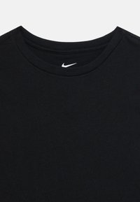 Melnā kreklā ar apaļu kakla izgriezumu, uz tā ir neliels balts Nike swoosh logo, kas izvietots centrā zem apkakles.