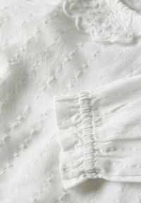 Weißer Baumwollstoff mit einer strukturierten Musterung, versehen mit gerafften Bündchen und einem floralen Akzent in der Nähe des Halsausschnitts. Detaillierte Nähte sind sichtbar.
