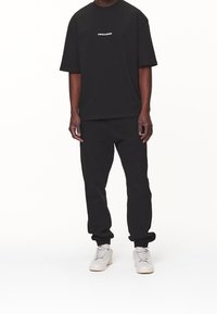 Svart bomullströja och byxset, avslappnad passform, med en vit logotyp på bröstet, i kombination med ljusgrå sneakers.