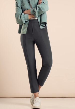 Pantalons gris extensibles avec une coupe ajustée, présentant une texture lisse et des coutures verticales. Associés à un chemisier vert clair et des baskets blanches.