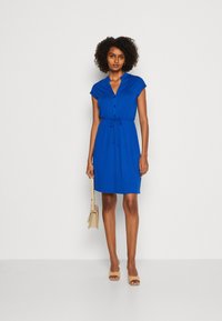 Vestito blu con bottoni, vita regolabile, maniche corte e una vestibilità comoda, abbinato a una borsa beige e scarpe aperte color terra.