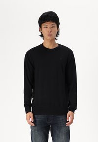 Schwarzer Pullover mit langen Ärmeln aus Strickstoff, mit rundem Ausschnitt und dezentem Logo-Detail. Getragen mit dunkelblauen Jeans.
