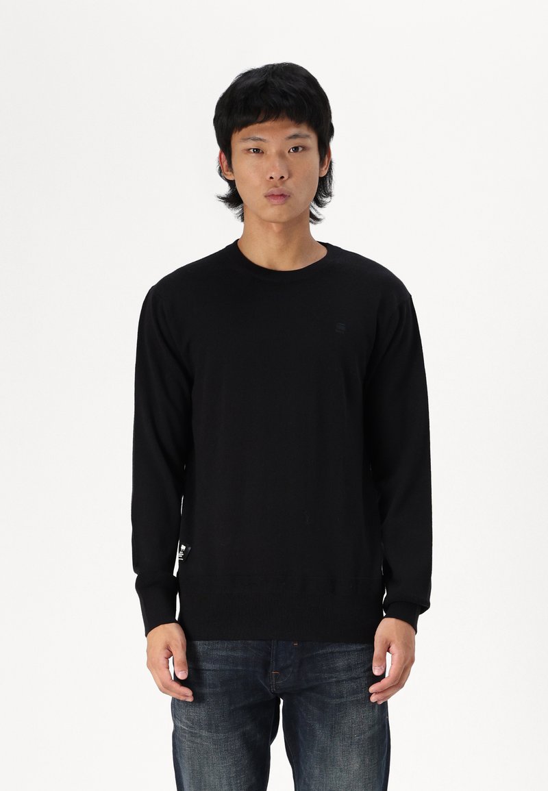 Schwarzer Pullover mit langen Ärmeln aus Strickstoff, mit rundem Ausschnitt und dezentem Logo-Detail. Getragen mit dunkelblauen Jeans.