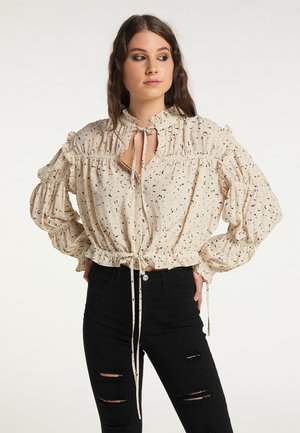 Junge Frau mit langen Haaren, die eine beige Bluse mit Rüschen und Bändern sowie schwarze zerrissene Skinny-Jeans trägt, steht mit den Händen in den Hüften.