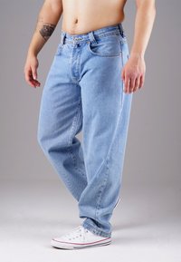Light blue denim jeans with a relaxed fit, featuring five pockets, contrasting orange stitching, and worn-in texture. Paired with white sneakers.