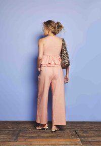 Hajadon, göndör hajjal, barackszínű ujjatlan, fodros felsőben és széles nadrágban, fehér szandált viselve, és egy leopárdmintás válltáskával a vállán áll, fapadlón.