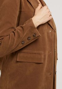 Veste en daim marron avec un col cranté, une poche poitrine simple et trois boutons marron au poignet. Texture lisse avec une coupe ajustée.