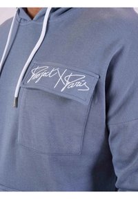 Hoody in gestructureerde lichtblauwe stof met een voorzak. Voorzien van witte trekkoorden en een geborduurd logo in cursief op de zak.