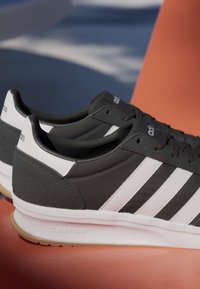 Zapatillas Adidas en blanco y negro con tres rayas blancas, entresuelas Cloudfoam y suelas de goma, colocadas sobre una superficie naranja.