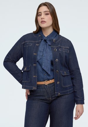 Kobieta w ciemnej kurtce dżinsowej, dopasowanej koszuli dżinsowej z kokardą, ciemnych dżinsach i beżowym pasku na jednolitym tle.