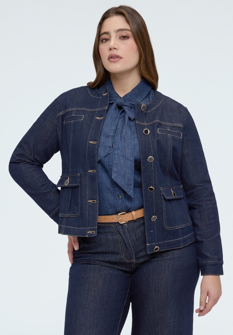 Kobieta w ciemnej kurtce dżinsowej, dopasowanej koszuli dżinsowej z kokardą, ciemnych dżinsach i beżowym pasku na jednolitym tle.