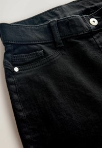 Close-up of black denim jeans showing waistband, belt loop, front pocket with small metal rivet, and white button on the waistband.