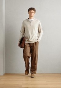 Suéter beige con cuello de cremallera, pantalones sueltos marrones y zapatos marrones. Modelo llevando un bolso marrón. Suelo liso con textura de madera clara. Fondo neutro.