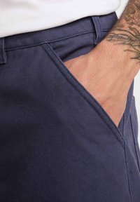 Temno modri kratki hlače iz teksturirane tkanine, s stranskimi žepi z vidnim šivom, v katerega je položena roka.