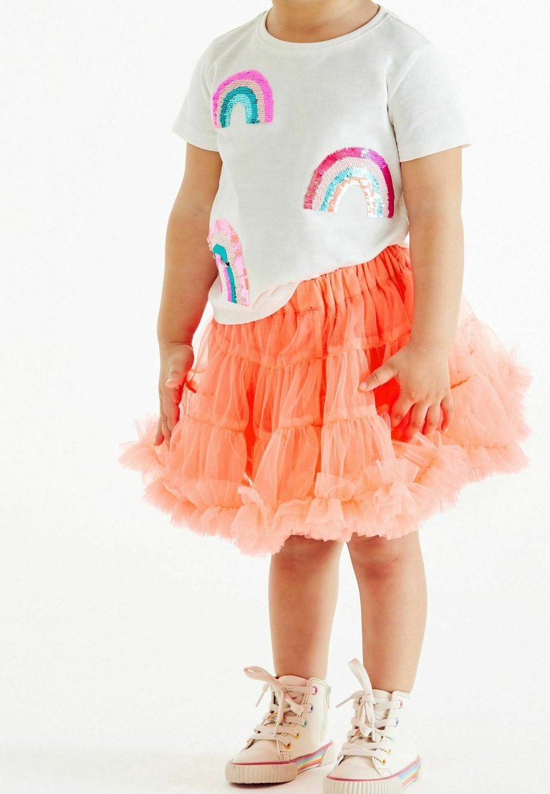 Tout-petit portant un t-shirt blanc avec des patchs arc-en-ciel colorés, une jupe corail à volants et des baskets montantes blanches avec des détails arc-en-ciel.