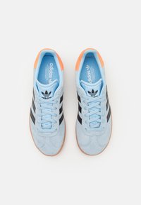 Light blue suede sneakers with black stripes, orange accents on the heel, and blue inner lining. Features a lace-up design and gum sole.