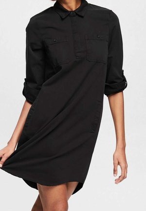 Femme portant une robe chemise noire à manches longues retroussées, avec des poches poitrine avant et un col, tenant l'ourlet de la robe.