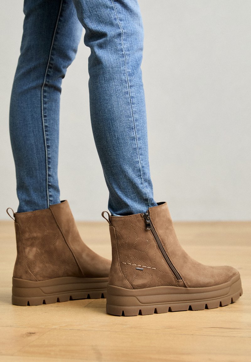Botas de ante marrón hasta el tobillo con un cierre lateral, diseño texturizado y una suela de goma gruesa con un patrón de tacos resistente. Combinadas con jeans azules.