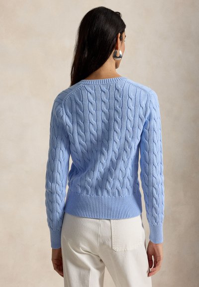 Hosszú, sötét hajú nő világoskék, kötött mintás pulóverben és fehér nadrágban, háttal áll a nézőnek.