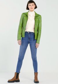 Groene leren jas met lange mouwen en ritszakken, gecombineerd met een lichtgekleurde blouse en blauwe strakke jeans; bruine enkellaarzen.
