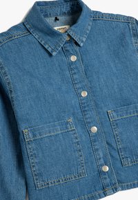 Denimjacka med krage, prydd med silverfärgad tryckknappar, två framfickor i patchstil och kontrasterande gul sömnad på en blå bakgrund.