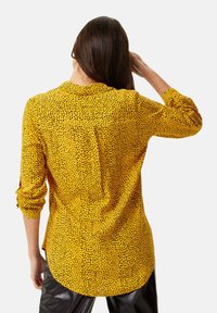 Yellow shirt featuring a dotted pattern, button-up front, long sleeves with rolled cuffs, and a curved hem at the back.