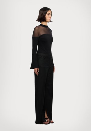 Black long-sleeve top with sheer mesh neckline, paired with high-waisted black trousers featuring slits at the cuffs, and strappy black sandals.