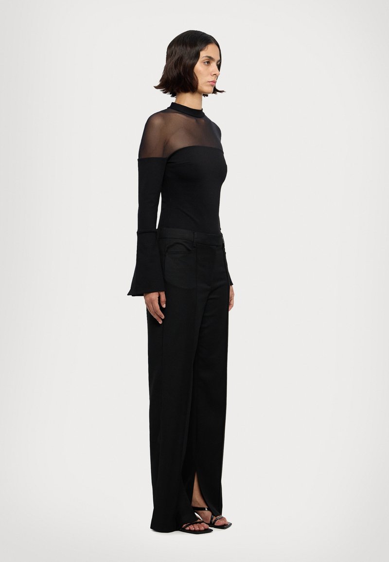 Haut noir à manches longues avec un col en tulle transparent, associé à un pantalon noir taille haute avec des fentes au niveau des chevilles, et des sandales noires à lanières.