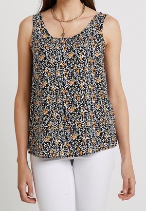 Kobieta w czarnym topie bez rękawów z drobnym białym i pomarańczowym kwiatowym wzorem, w połączeniu z białymi spodniami i złotymi naszyjnikami łańcuszkowymi.