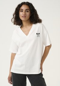 T-shirt bianco in cotone con scollatura a V e maniche corte, caratterizzato da un piccolo design con fiocco nero sul lato sinistro del petto. Tessuto morbido, vestibilità rilassata.