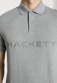Polo gris avec un col classique, présentant deux boutons et un logo "HACKETT" bien visible en gris foncé sur la poitrine.