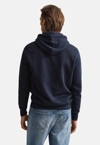 Sweat à capuche bleu marine à coupe décontractée, avec capuche à cordon et poignets côtelés. Tissu épais en mélange de coton avec une texture lisse. Porté avec un jean bleu clair.
